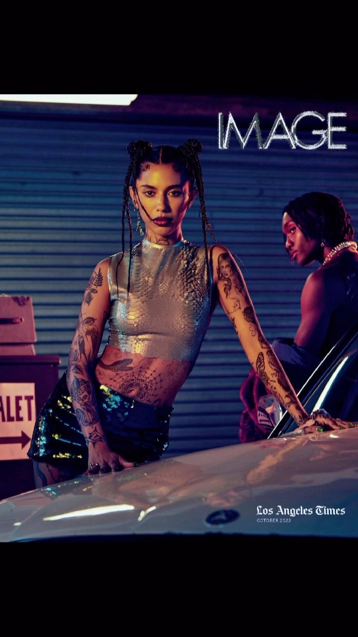Magazine cover featuring a woman with tattoos leaning on a car, with another person in the background, against a garage door backdrop.