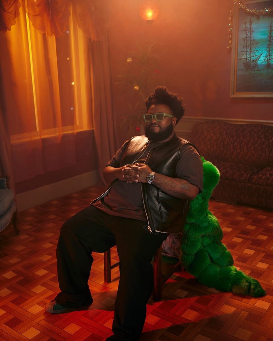 An adult male with beard and sunglasses sits indoors on a chair near a couch with a Christmas tree in the background.