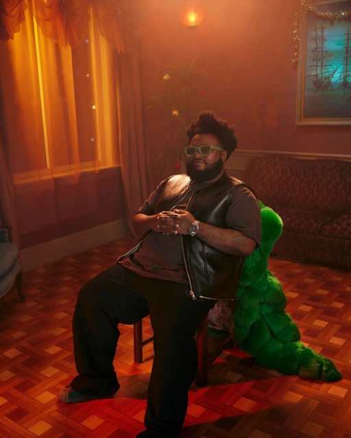 An adult male with beard and sunglasses sits indoors on a chair near a couch with a Christmas tree in the background.