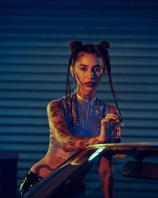 Portrait of a tattooed woman with braided hair, wearing a metallic top, resting her arm on a metal structure in an urban setting.