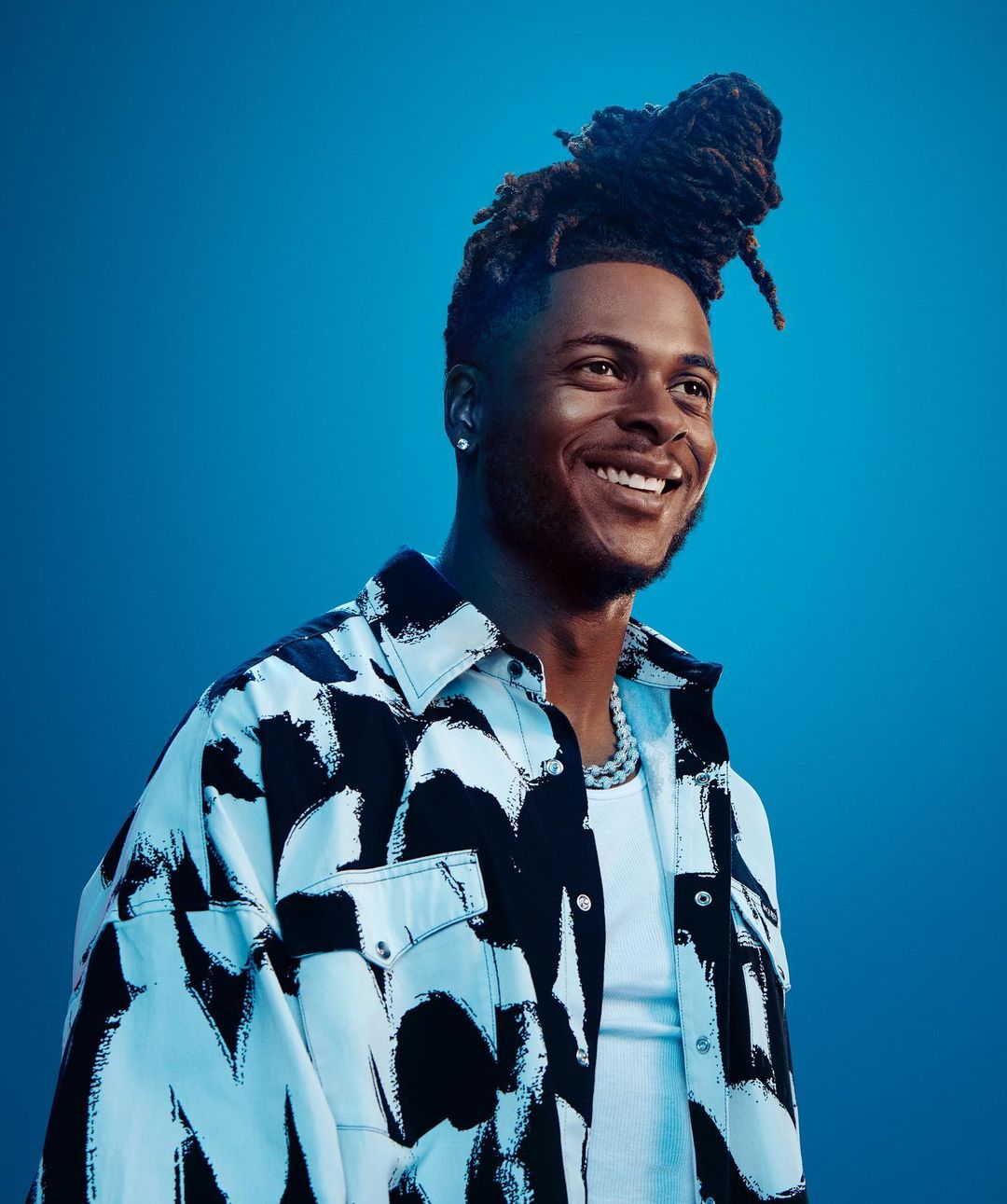 Portrait of an adult man with dreadlocks smiling in a patterned shirt against a blue background in a studio setting.