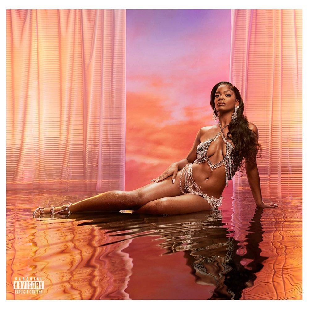 Glamorous adult woman poses in a shimmering crystal bikini and jewelry, reclined on reflective surface, against background of peach and lavender tones.