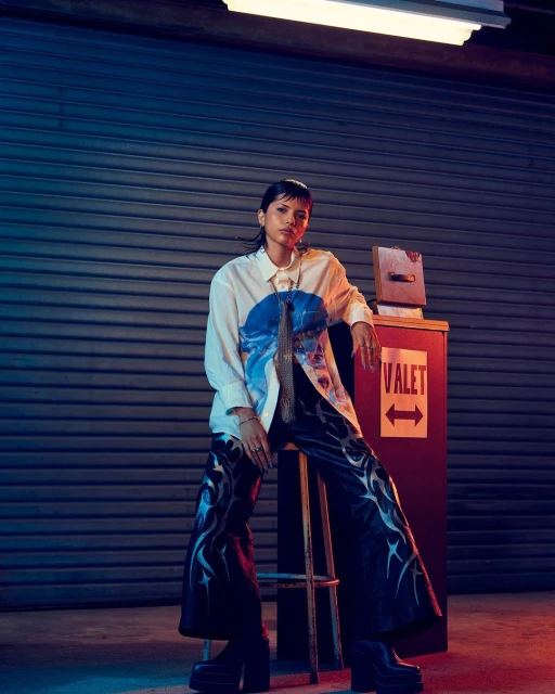 Stylish young woman poses in a white printed shirt and flame-printed pants, seated on a stool, in front of a metal garage door.