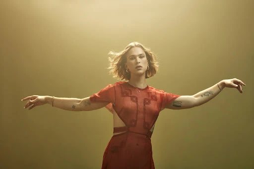 A woman with short blonde hair poses with outstretched arms in a red dress against a light-colored background in an eye-level studio shot.