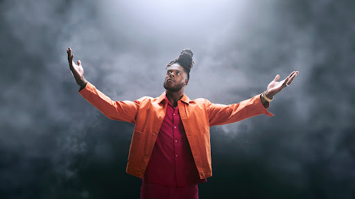 Black man with dreadlocks wearing an orange jacket and red shirt with arms raised against a dark smoky background.
