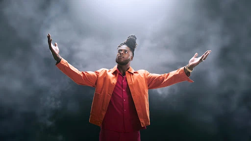 Black man with dreadlocks wearing an orange jacket and red shirt with arms raised against a dark smoky background.