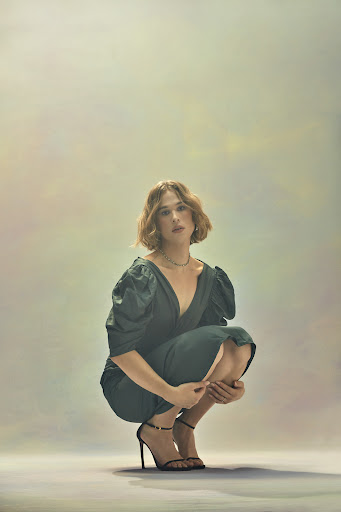 A poised woman with short brown hair, dressed in a dark green dress and strappy black heels, crouches against a mottled, neutral background.