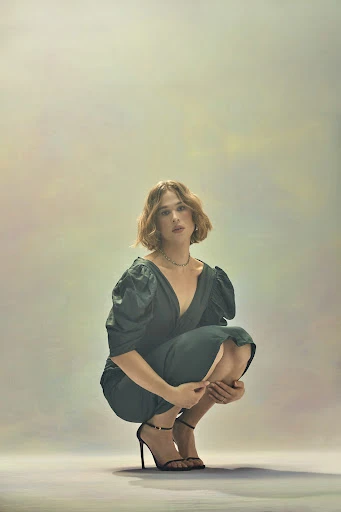 A poised woman with short brown hair, dressed in a dark green dress and strappy black heels, crouches against a mottled, neutral background.