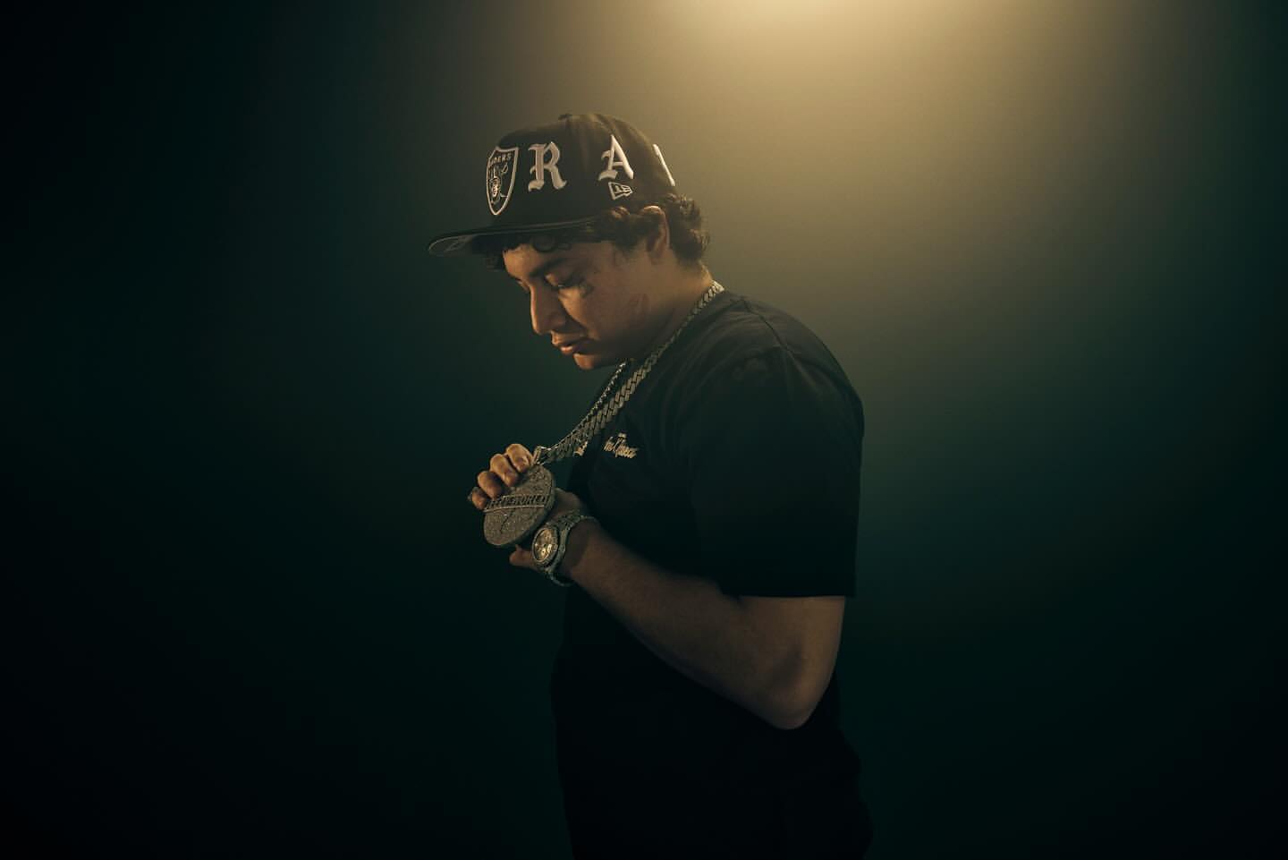 Young man wearing a black baseball cap and t-shirt, holding a large pendant necklace under dramatic overhead lighting by Gizelle Hernandez.