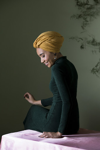 Woman wearing yellow turban and green dress sitting on a pink table in front of a green wall with dark smoky patterns.
