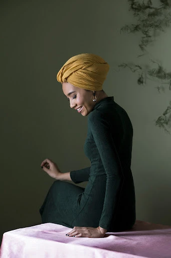 Woman wearing yellow turban and green dress sitting on a pink table in front of a green wall with dark smoky patterns.