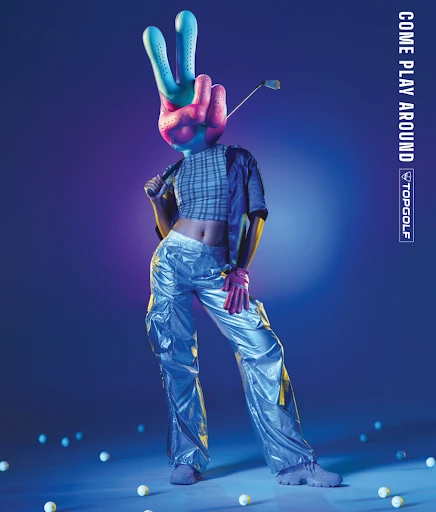 A person with a novelty peace-sign hand head poses in shiny silver pants, holding a golf club, with scattered golf balls below.