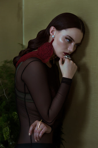 Elegant woman in sheer black top poses by wall with large tassel earring, jewelry, and greenery for fashion editorial.