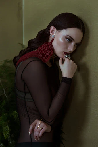 Elegant woman in sheer black top poses by wall with large tassel earring, jewelry, and greenery for fashion editorial.