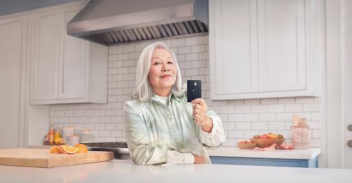 Elderly woman in kitchen holding a black card, smiling, with fruits and a cutting board on a bright countertop.