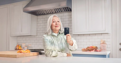 Elderly woman in kitchen holding a black card, smiling, with fruits and a cutting board on a bright countertop.