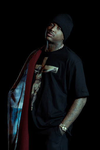 Portrait of a man in a black graphic t-shirt and beanie standing against a dark background, photographed by Gizelle Hernandez.