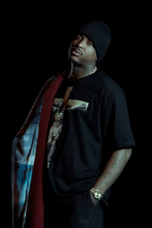 Portrait of a man in a black graphic t-shirt and beanie standing against a dark background, photographed by Gizelle Hernandez.