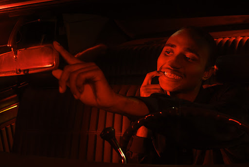 Young adult in a vintage car, bathed in red light, steering wheel visible, inside setting, driving, focus on the smiling male.