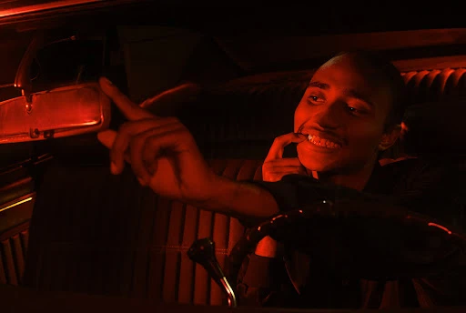 Young adult in a vintage car, bathed in red light, steering wheel visible, inside setting, driving, focus on the smiling male.