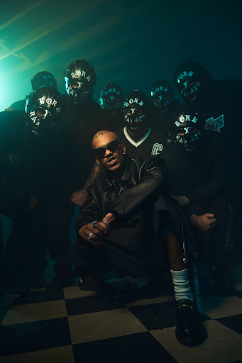 Male singer squats on a checkered floor with masked figures in the background against smoky green studio lighting for a music video.