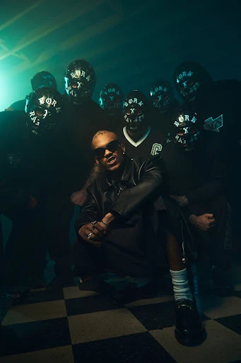 Male singer squats on a checkered floor with masked figures in the background against smoky green studio lighting for a music video.