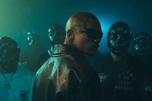 Close-up of a man with sunglasses and tattoos, leather jacket, surrounded by masked figures in a dimly lit indoor environment.