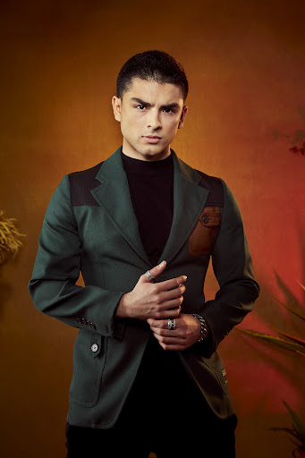 Portrait of a young man in a dark green blazer with leather details, a black turtleneck, rings, and a bracelet posing in a warm-toned studio.
