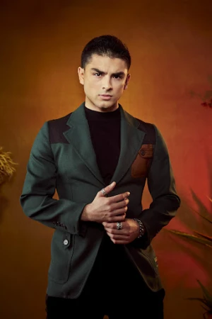 A man wearing a green blazer and black turtleneck posing against a warm textured background, photographed by Gizelle Hernandez.
