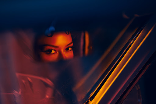 Close-up of a woman's face inside a car, illuminated by orange light and contrasted by deep blue tones, with focus on her expressive eyes.