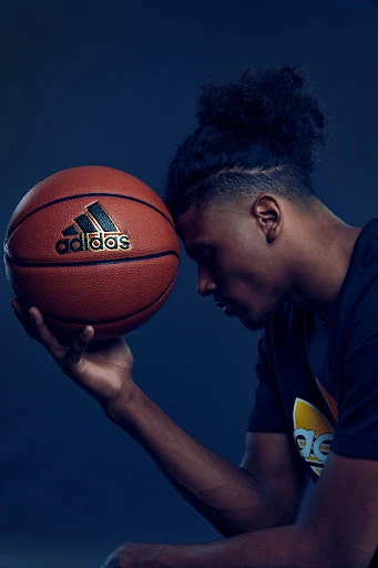 Portrait of Black male athlete holding Adidas basketball, close-up shot against a gradient blue background in a studio setting.