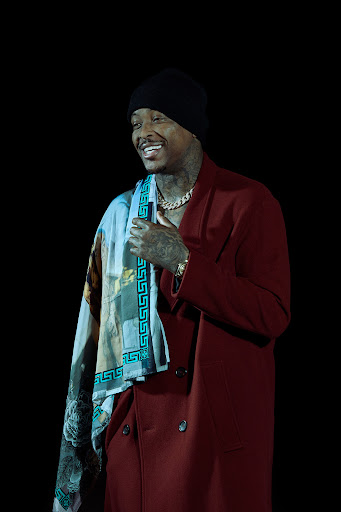 Adult male with tattoos smiles in a studio shot wearing a black beanie, burgundy suit jacket, gold chain, and patterned scarf.