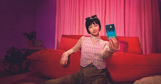 Young Asian woman sits indoors with a modern aesthetic, holding a credit card with a stylized evergreen forest graphic on a teal background.