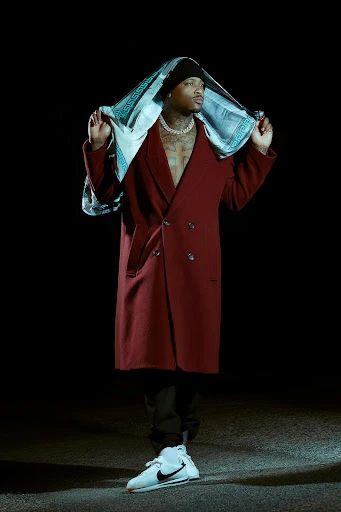 A man wearing a red coat and patterned head covering poses against a black background with a concrete floor in a studio setting.