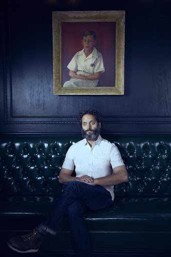 Adult male with beard sits on a green leather couch in an indoor space with dark walls and a portrait hanging above the couch.