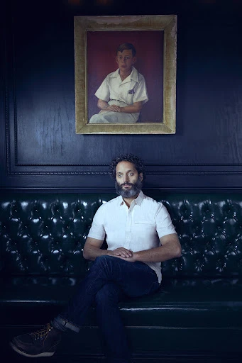 Adult male with beard sits on a green leather couch in an indoor space with dark walls and a portrait hanging above the couch.