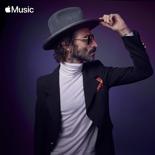Portrait of an adult male with a beard and sunglasses in a black suit and grey hat against a purple background, artistic and stylish.