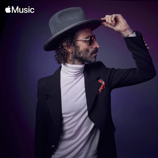 Portrait of an adult male with a beard and sunglasses in a black suit and grey hat against a purple background, artistic and stylish.