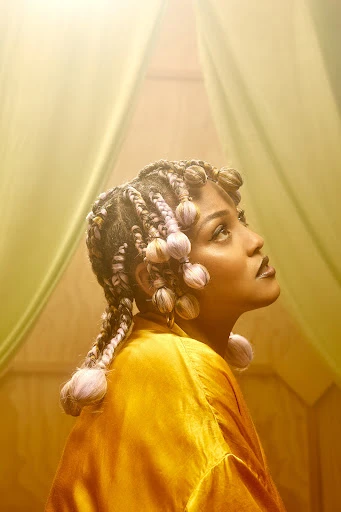 Portrait of a woman with braided hair and a yellow robe looking up in a warm, brightly lit indoor studio with muted green curtains.