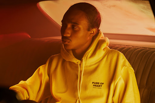 Young Black man in yellow hoodie inside vintage car with plush interior and moody, low lighting for fashion or entertainment content.