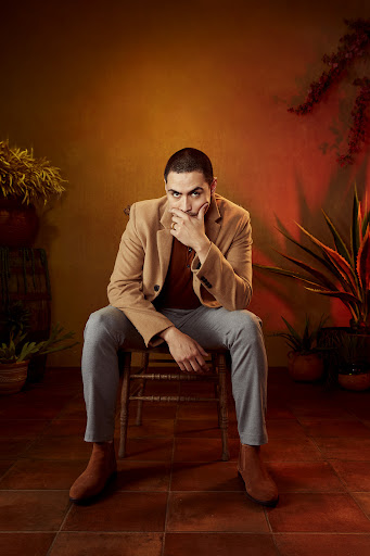 Portrait of an adult male with a hand on his face, wearing a tan blazer and blue jeans, sitting on a wooden chair indoors.
