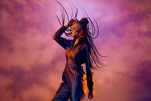 Smiling woman with braided hair wearing a black jumpsuit against a textured purple and orange background with cloud-like formations.