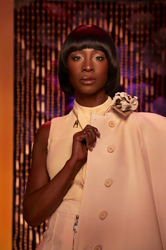 Elegant dark-skinned woman with a sleek bob hairstyle poses in front of a decorative beaded curtain in a sleeveless top and trousers.