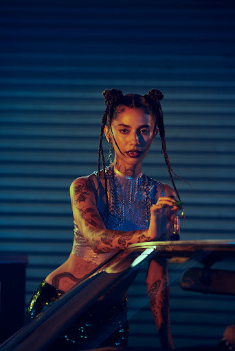 Portrait of a tattooed adult female with braids and an expressive face, wearing a shiny silver top, posing indoors against blue background.