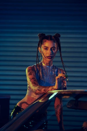 Portrait of a tattooed adult female with braids and an expressive face, wearing a shiny silver top, posing indoors against blue background.