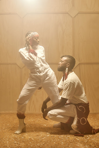 Two models strike poses in a studio illuminated with soft, diffused lighting; the woman in a jumpsuit and the man in casual clothes.