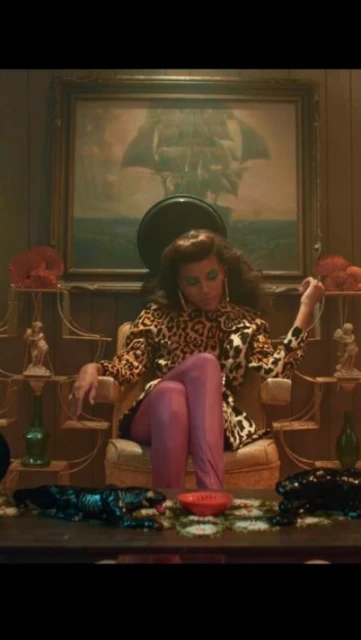 Woman in a leopard-print jacket sits at a desk with decorative animal sculptures, a painting of a ship, and vintage accessories.