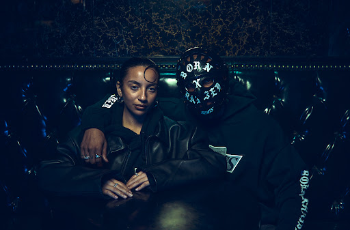 A woman and a person in a mask pose indoors. The woman wears a jacket and has jewelry, and the masked person wears a hoodie.