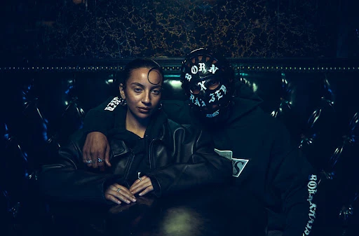A woman and a person in a mask pose indoors. The woman wears a jacket and has jewelry, and the masked person wears a hoodie.