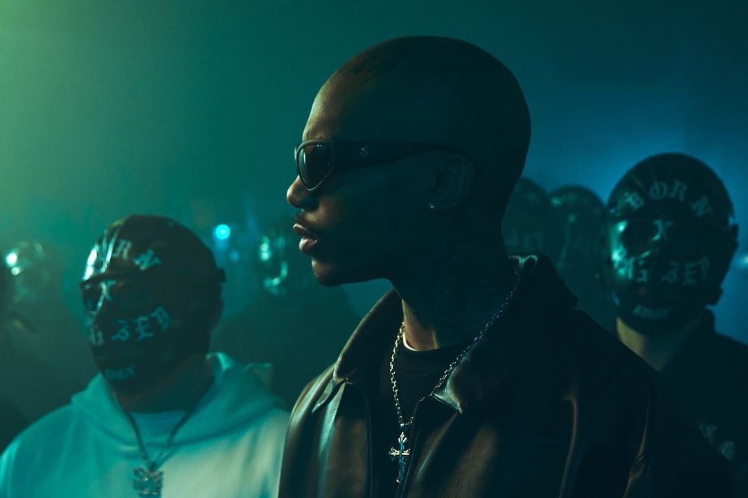 Close up of an adult male wearing sunglasses and a chain, surrounded by a crowd with masks, against a blue-green background.